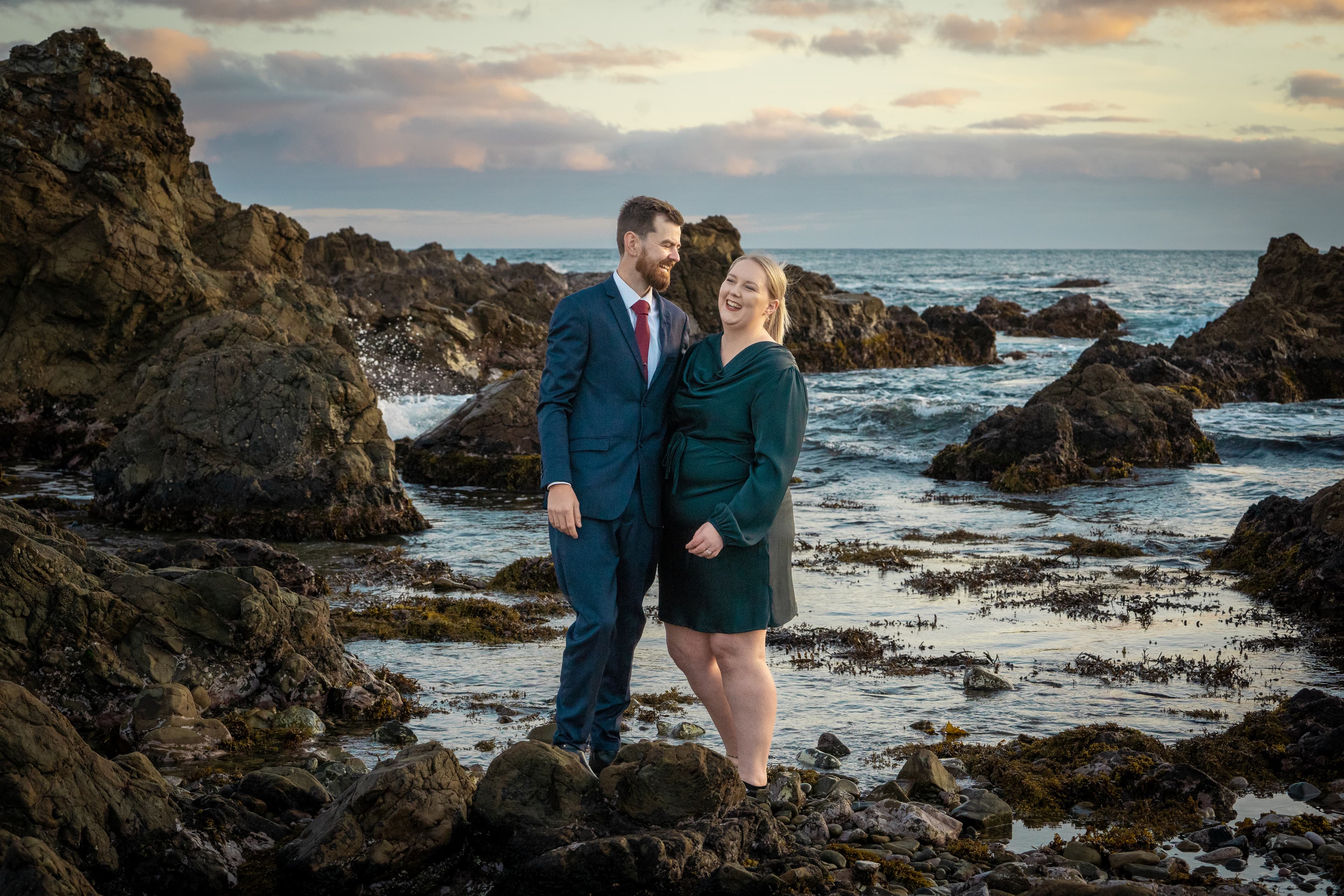 Man in blue suit and woman in green dress laugh on a rocky beach.